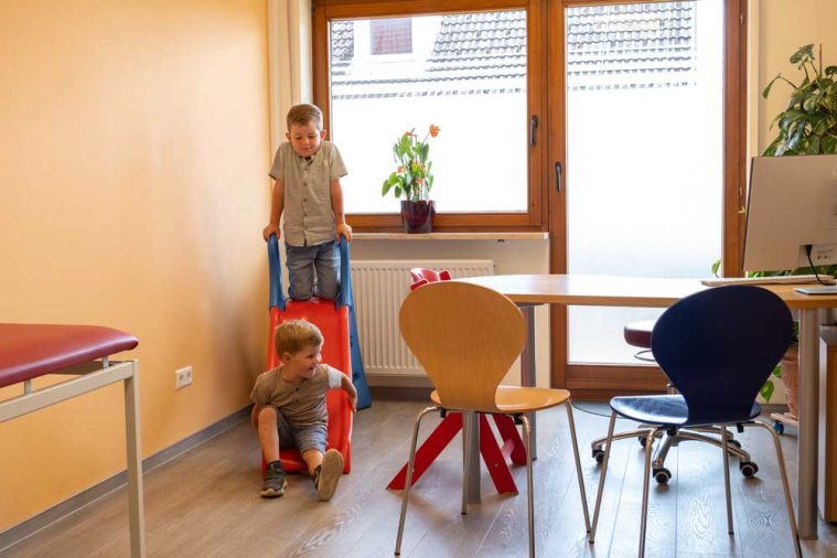 Zwei Kinder spielen mit einem roten Wagen in einem hellen Raum mit Pflanzen.